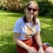 Chloe sat on the grass smiling at the camera. She is wearing sunglasses, a white top and an orange skirt