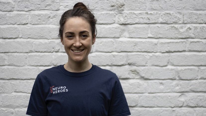 Bex, woman with brown hair in a bun. She’s stood against a white wall and is wearing a navy t-shirt