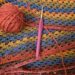 Ball of orange wool and a purple crochet hook. The background is a large crocheted blanket