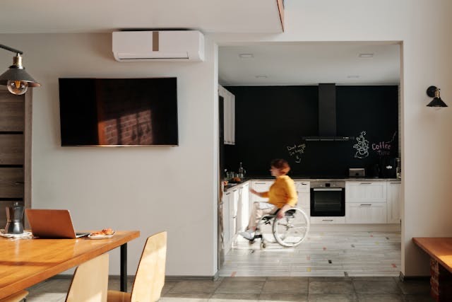 A woman in a grey and wood kitchen. She is using a silver wheelchair and is wearing a yellow jumper